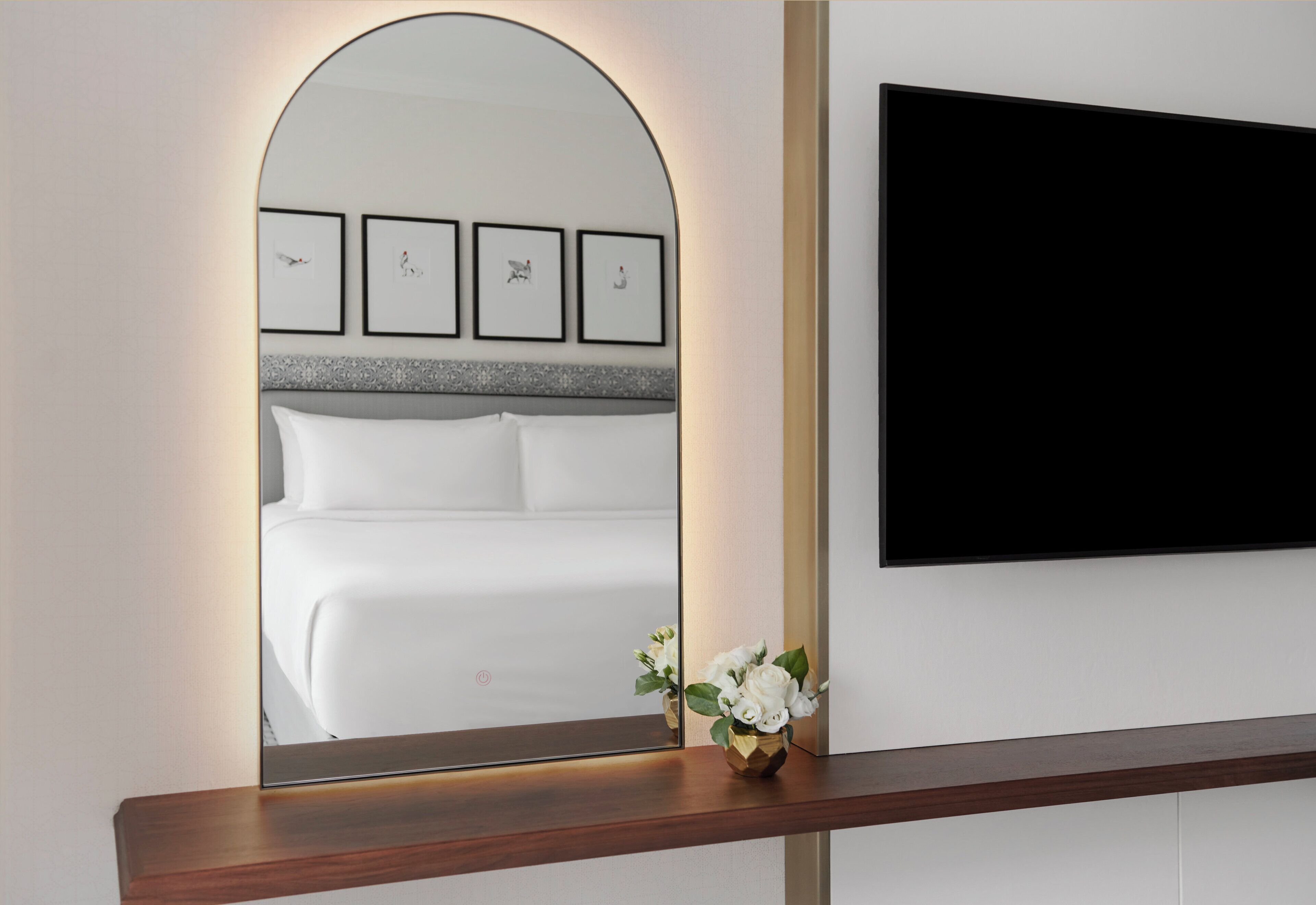 Habitación de hotel con un espejo grande que refleja una cama, un televisor de pantalla plana montado en la pared y un pequeño jarrón con flores sobre una estantería de madera.
