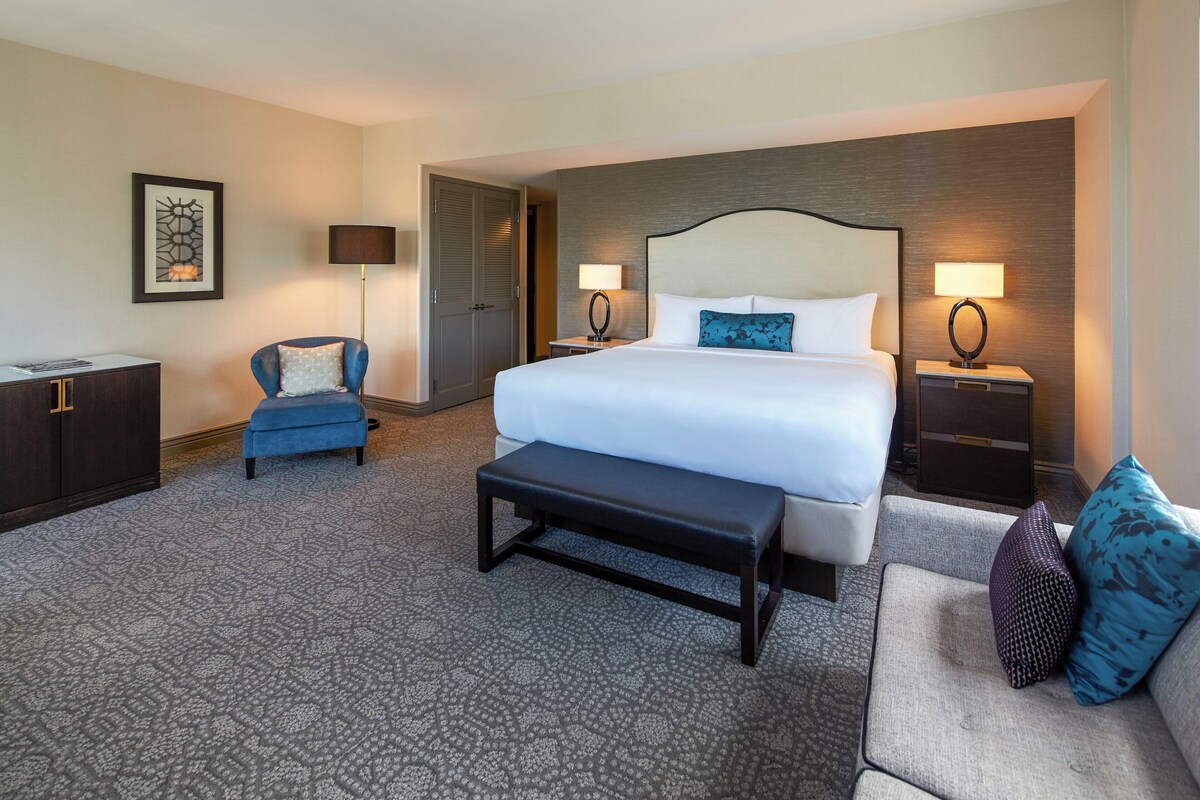 A hotel room with a large bed, a bench, a chair, a desk, and two bedside tables with lamps.
