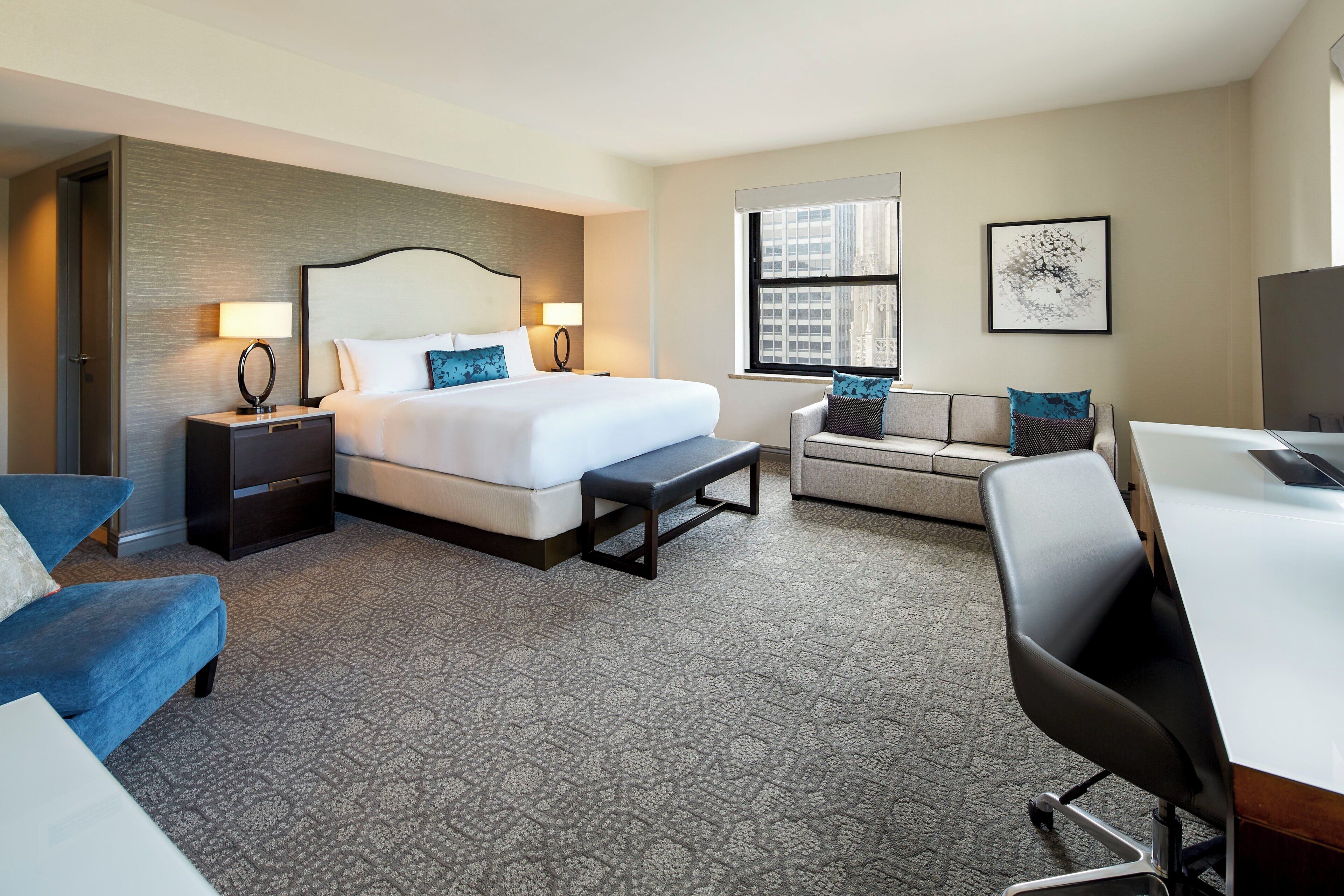 A hotel room with a bed, a desk, a chair, a sofa, a bench, a nightstand, and a window.