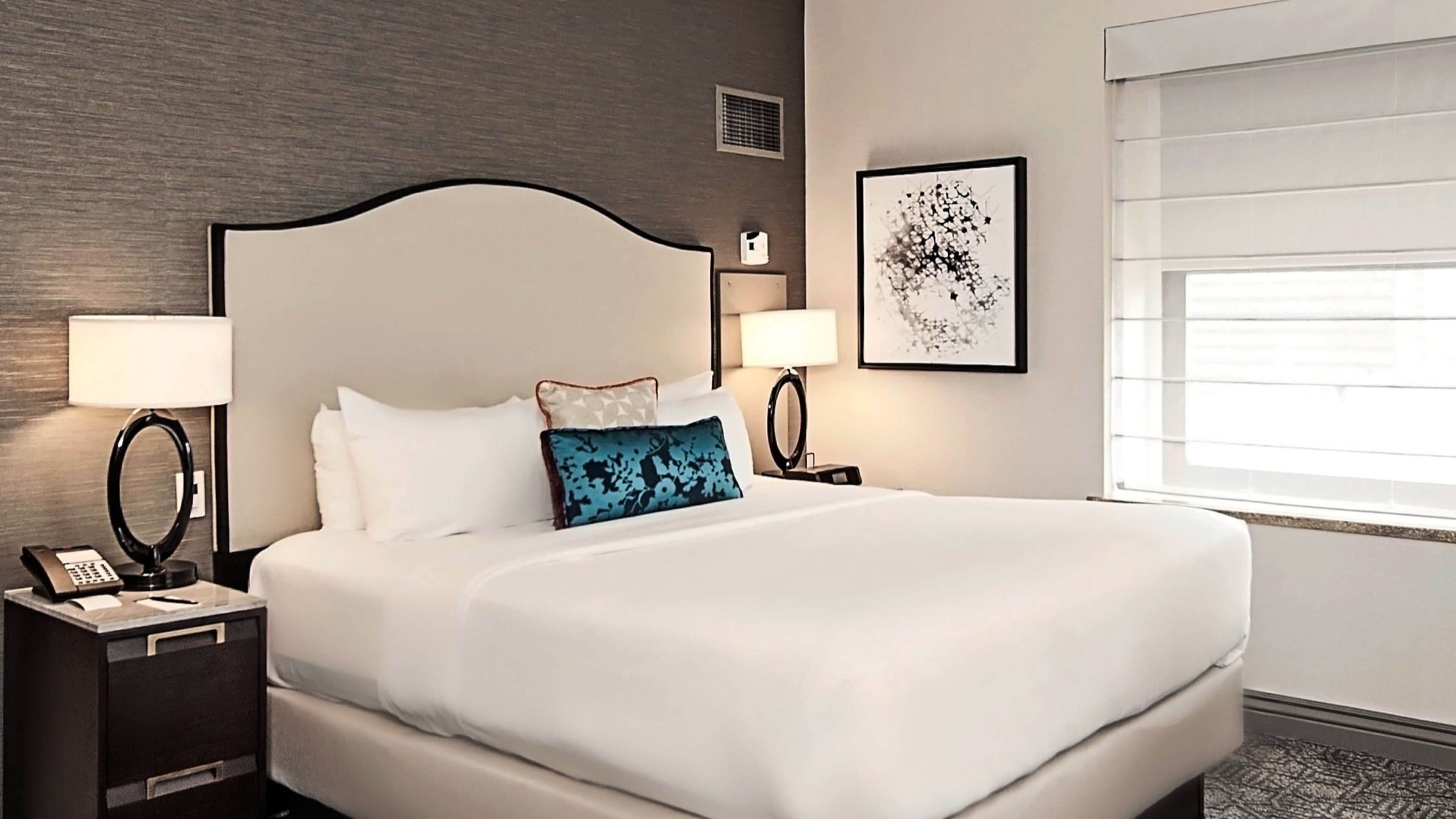Habitación de hotel con una cama grande, dos mesitas de noche con lámparas, una obra de arte en la pared y una ventana con persianas.