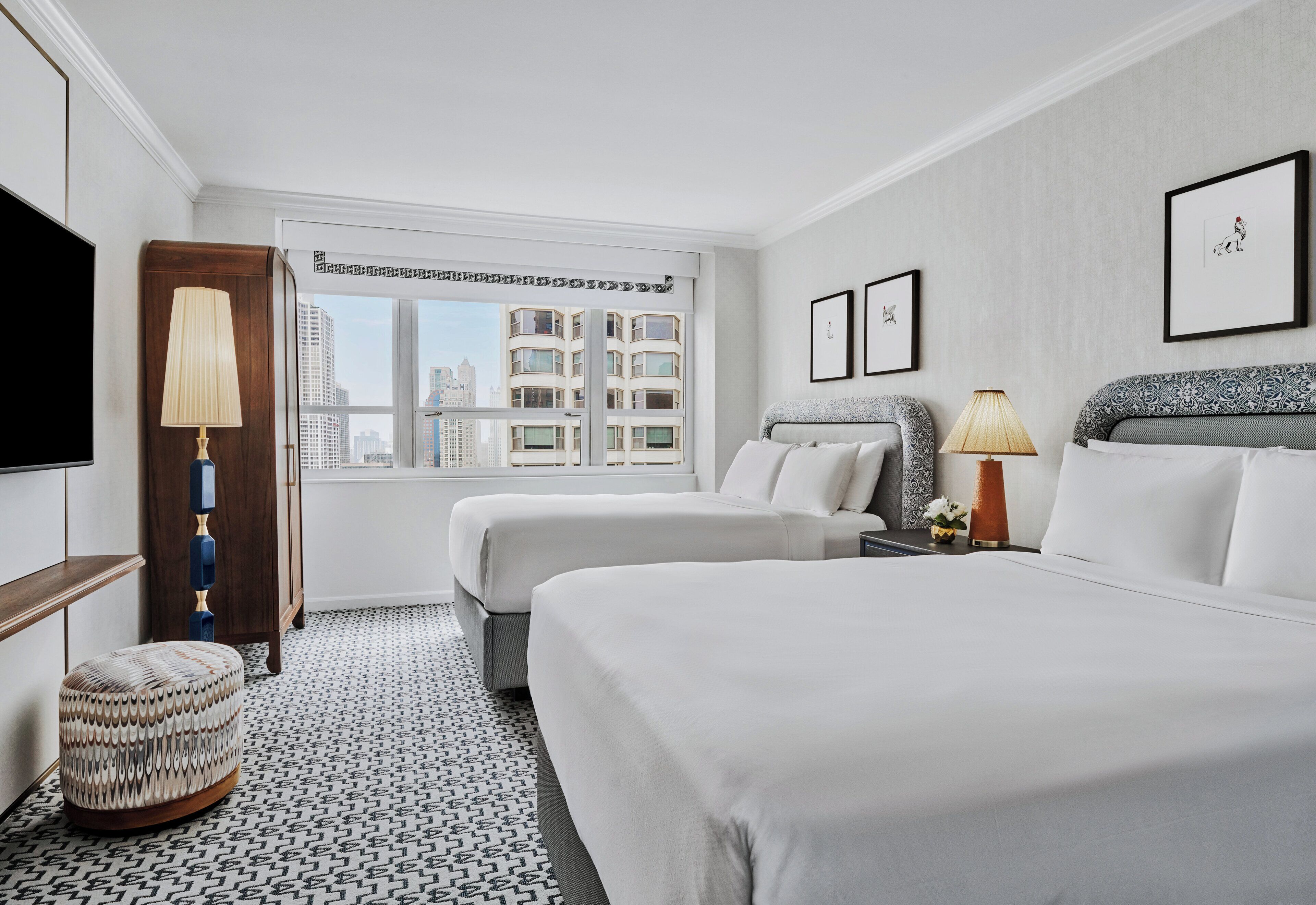 A hotel room with two beds, a window with a city view, a TV, and a lamp.