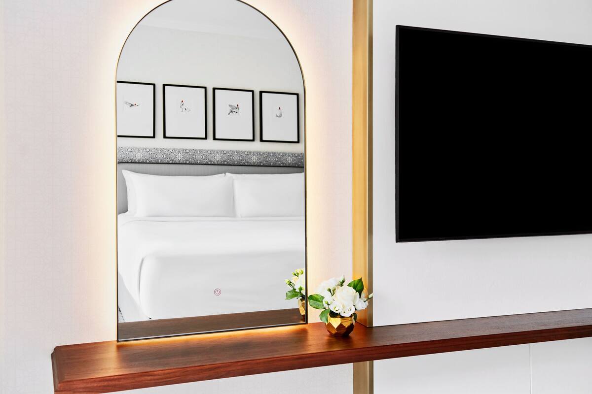 Un dormitorio con una cama, una mesita de noche con un jarrón de flores y un espejo grande que refleja la cama y las obras de arte en la pared.