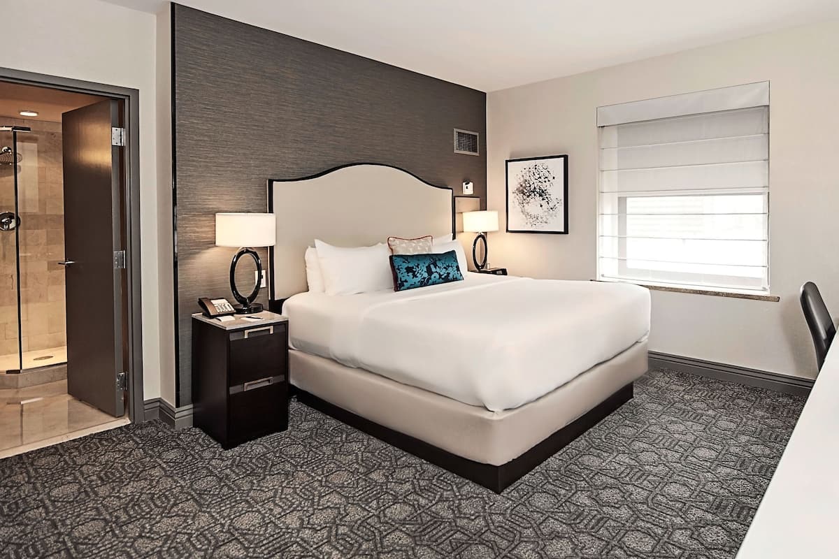 Habitación de hotel con una cama grande, un escritorio con silla, una ventana con persianas, una puerta y un área de ducha.