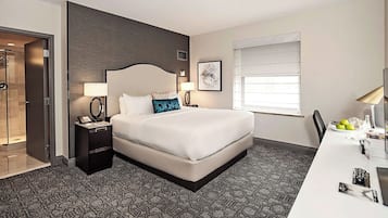 A hotel room with a large bed, bedside tables with lamps, a desk with a chair, a window with blinds, and a door leading to a shower area.