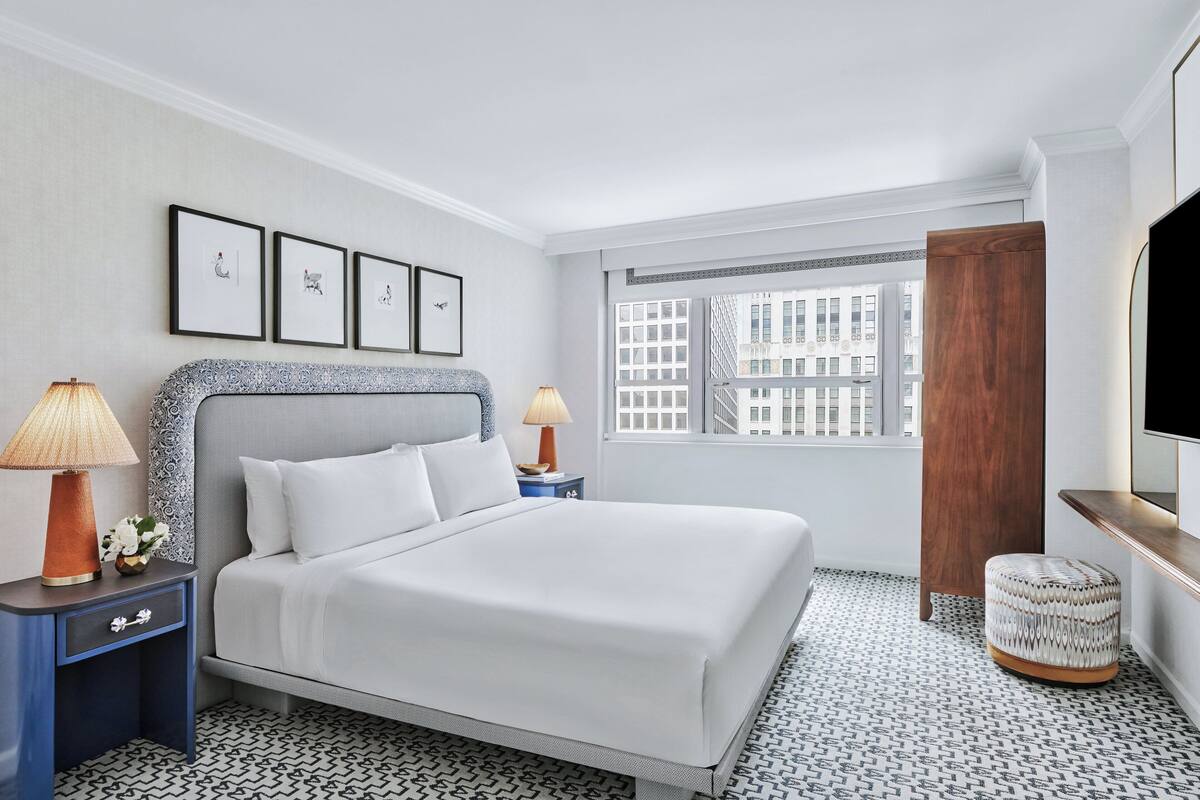 Habitación de hotel con una cama grande, dos lámparas de noche, una mesita con lámpara, televisión y ventana con vista a la ciudad.
