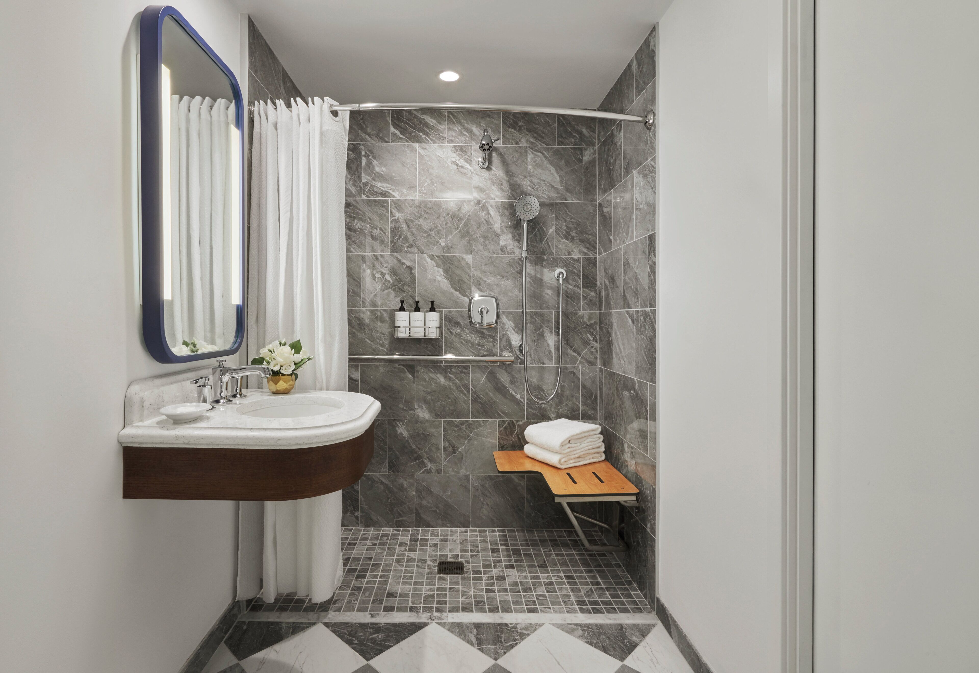 A modern bathroom with a marble shower, a sink with a mirror, and a small wooden shelf with towels.