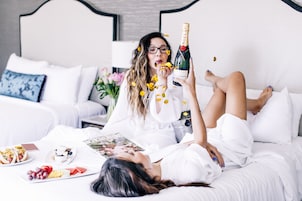 Una persona recostada en una cama con una botella de champán, rodeada de comida y una revista.