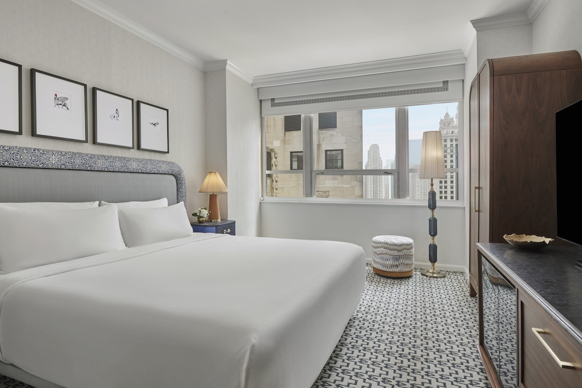 A hotel room with a large bed, a nightstand with a lamp, a small round table, and a view of the city through a window.