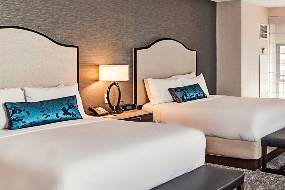A hotel room with two beds, a nightstand with a lamp, and a small table.