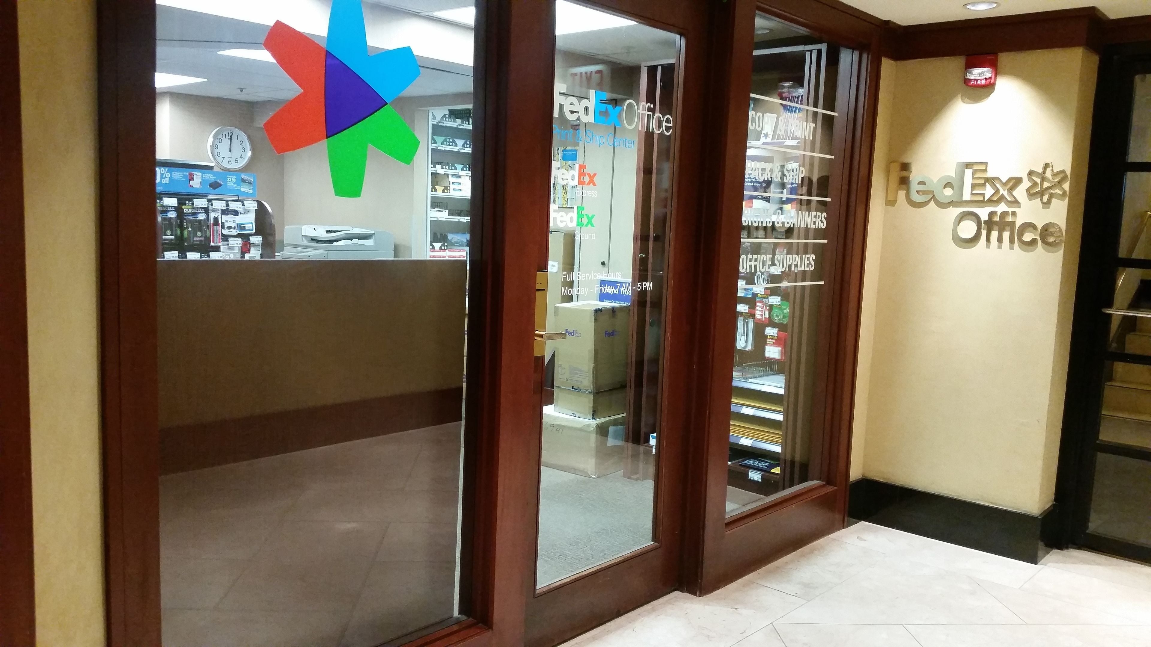 A FedEx Office with a large logo on the wall, a clock, and various office supplies visible through the glass doors.