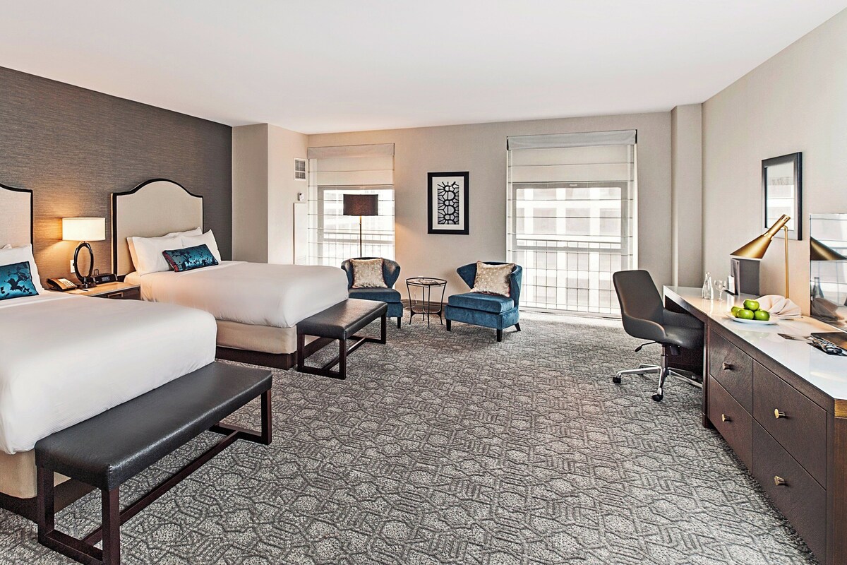 A hotel room with two beds, a desk, a chair, a small table, and a window.