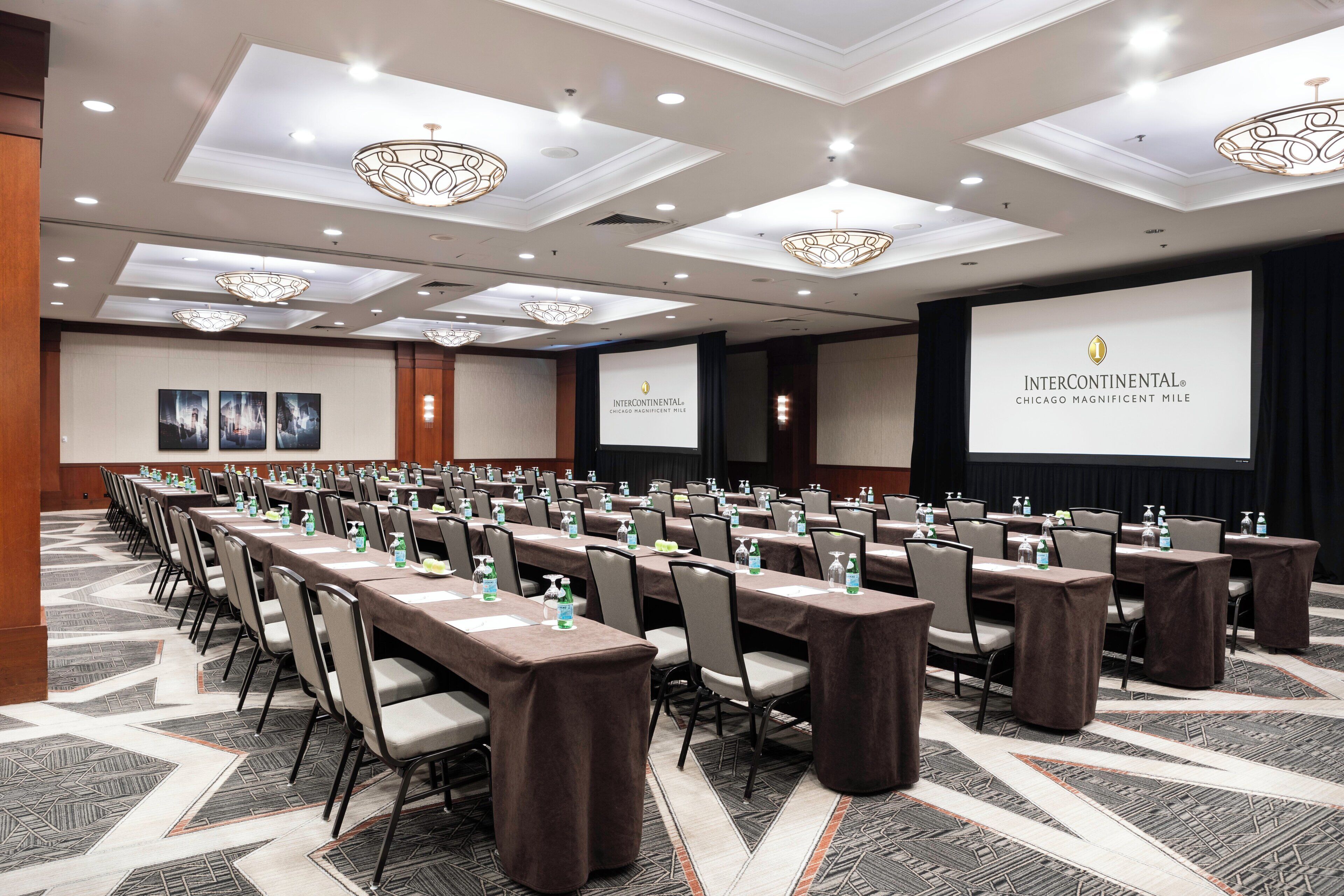 Un salón de conferencias con mesas largas, sillas y una pantalla grande que muestra el logotipo de InterContinental Chicago Magnificent Mile.