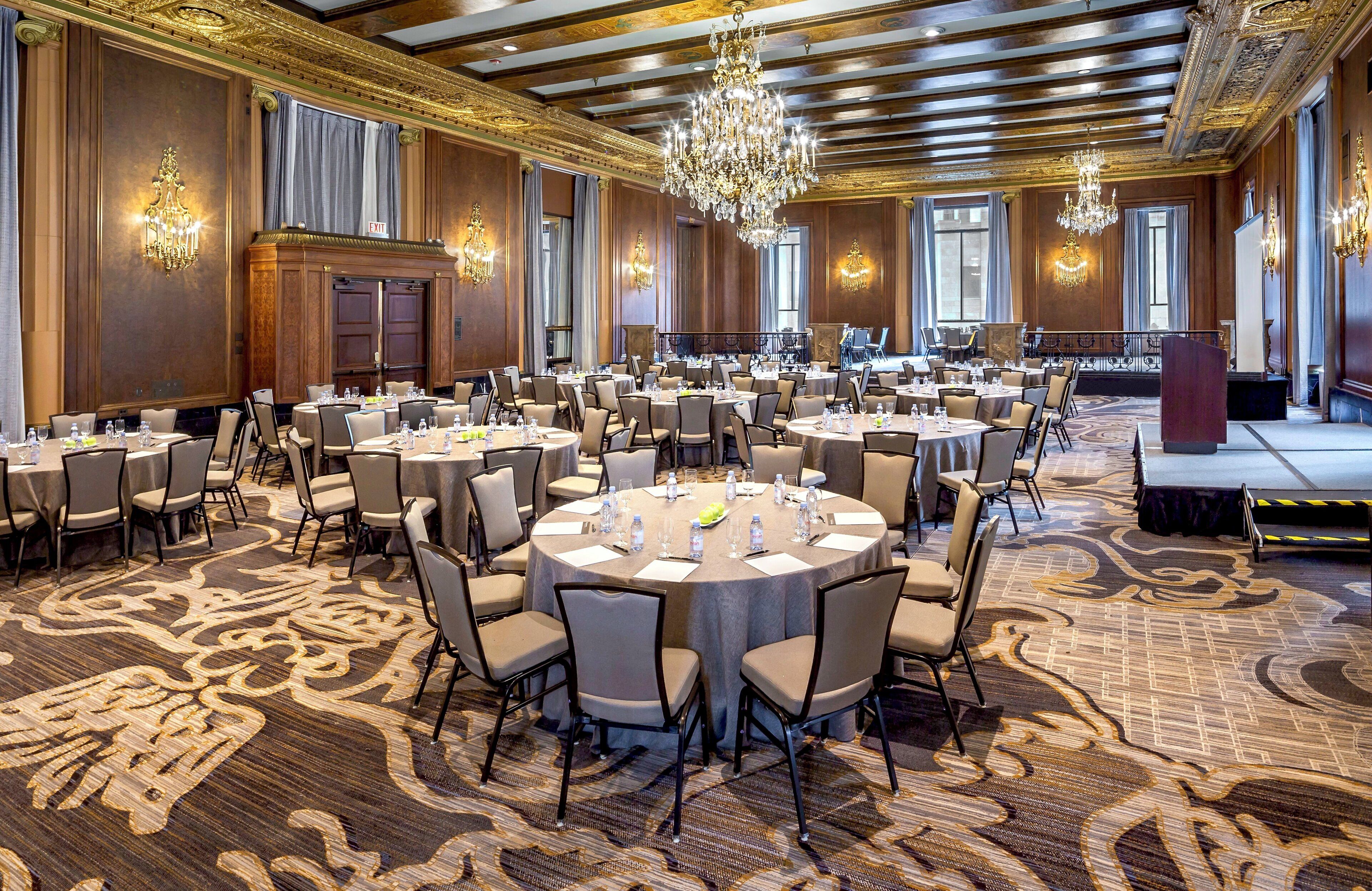 A large banquet hall with round tables, chairs, chandeliers, and a patterned carpet.