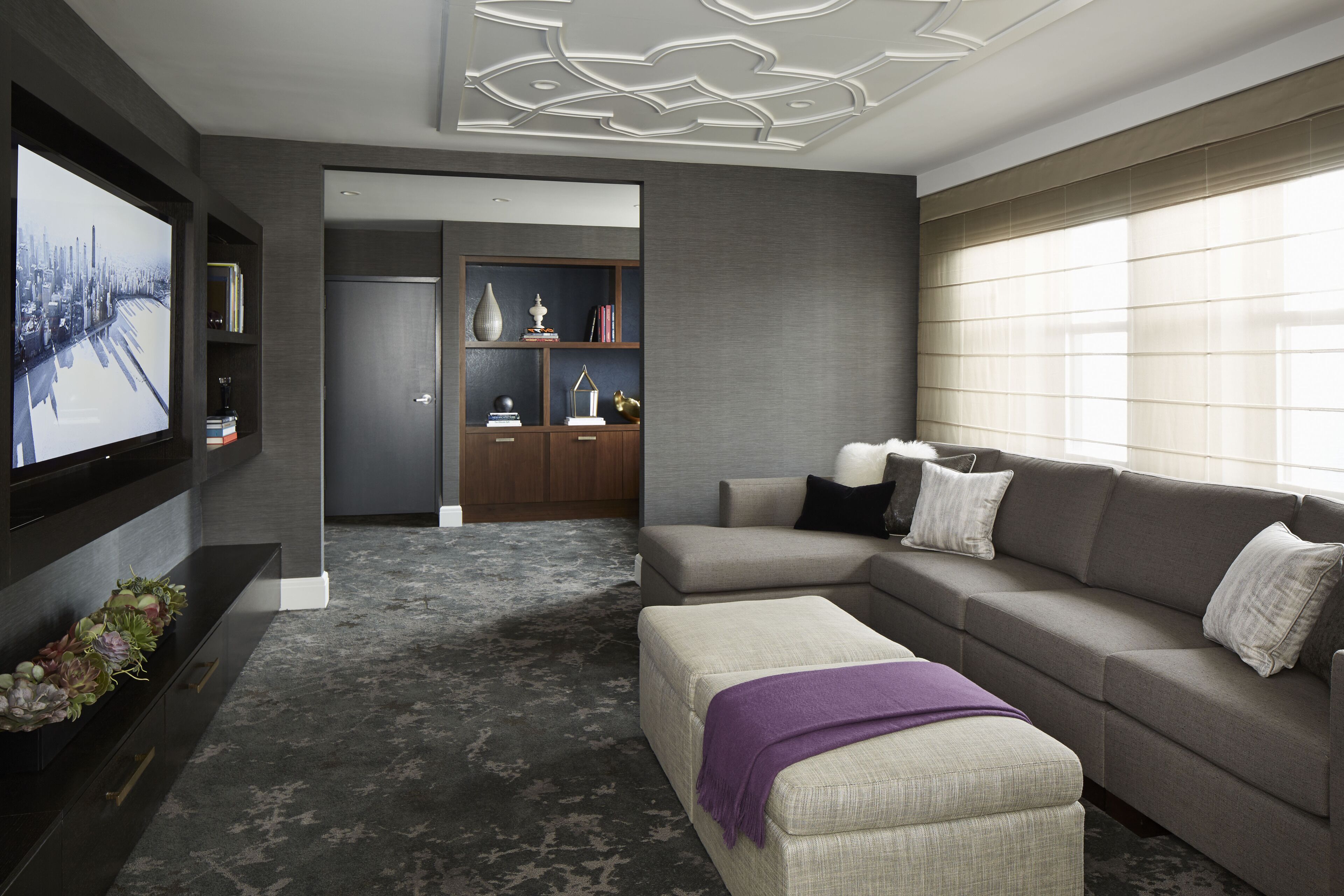 A modern living room with a sectional sofa, a flat-screen TV mounted on the wall, a bookshelf, and a decorative ceiling.