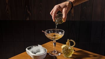 Une main verse une boisson dans un verre posé sur une table en bois, à côté d’un bol de glaçons et d’une bouteille de spiritueux.