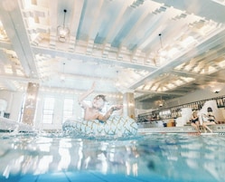 A swimming pool area with a glass-bottomed pool, a high ceiling with hanging lights, and a staircase with people sitting on it.