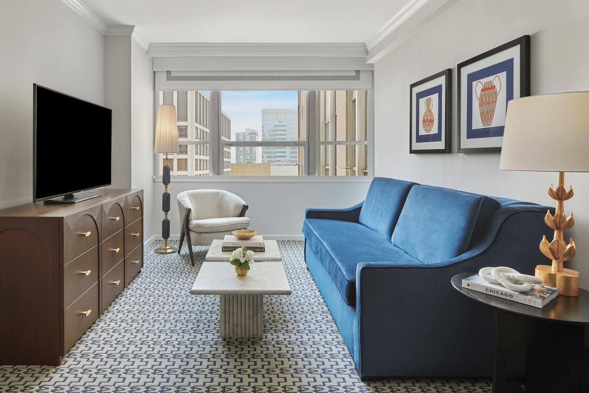 Un salón moderno con un sofá azul, un televisor de pantalla plana, una mesa de centro y vistas a la ciudad a través de amplias ventanas.