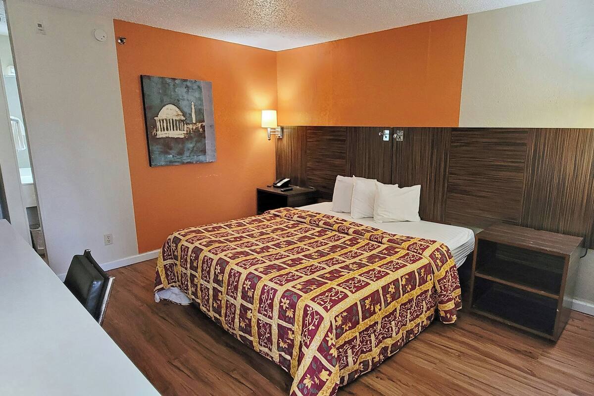 Ein Hotelzimmer mit Holzboden, einem Bett mit gemustertem Überwurf, einem Nachttisch mit Lampe sowie einem Bild an der Wand.