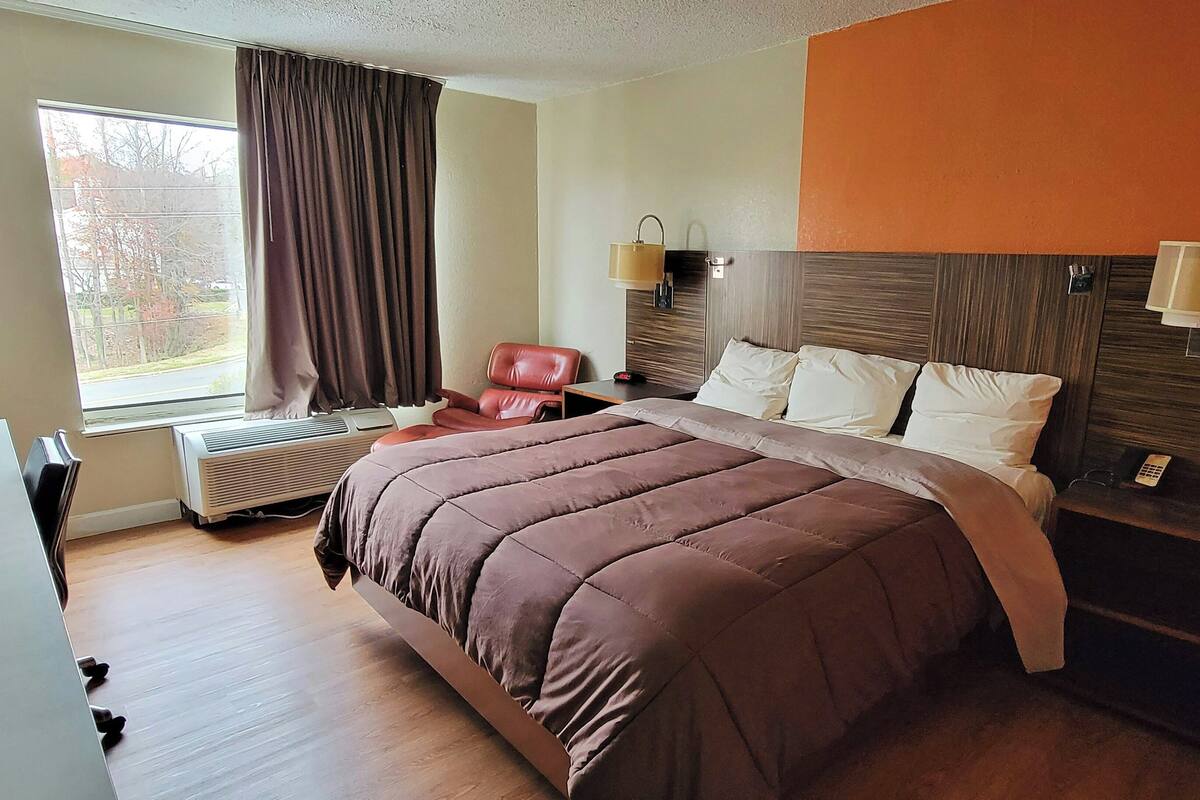 Ein Hotelzimmer mit einem Bett, einem Schreibtisch, einem Stuhl, einem Fenster mit Vorhängen und einer Wandlampe.