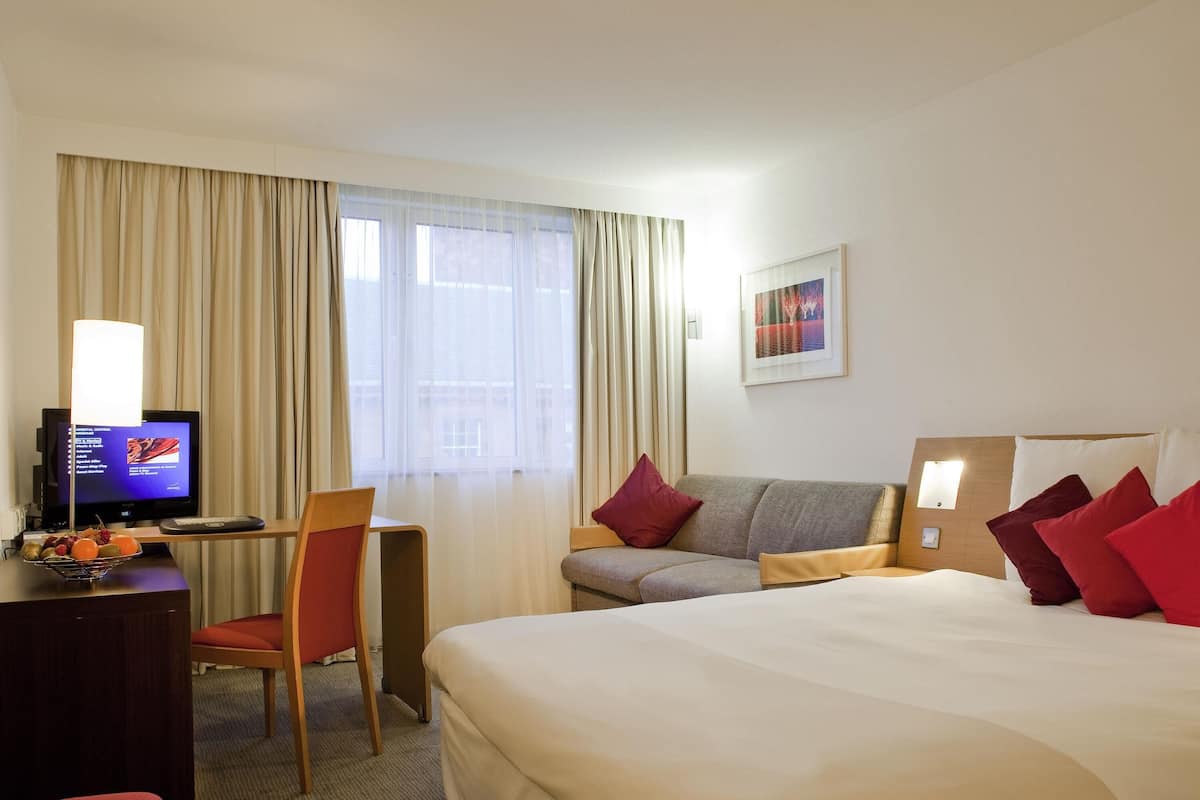 A hotel room with a bed, a desk with a computer, a chair, a television, and a window with curtains.