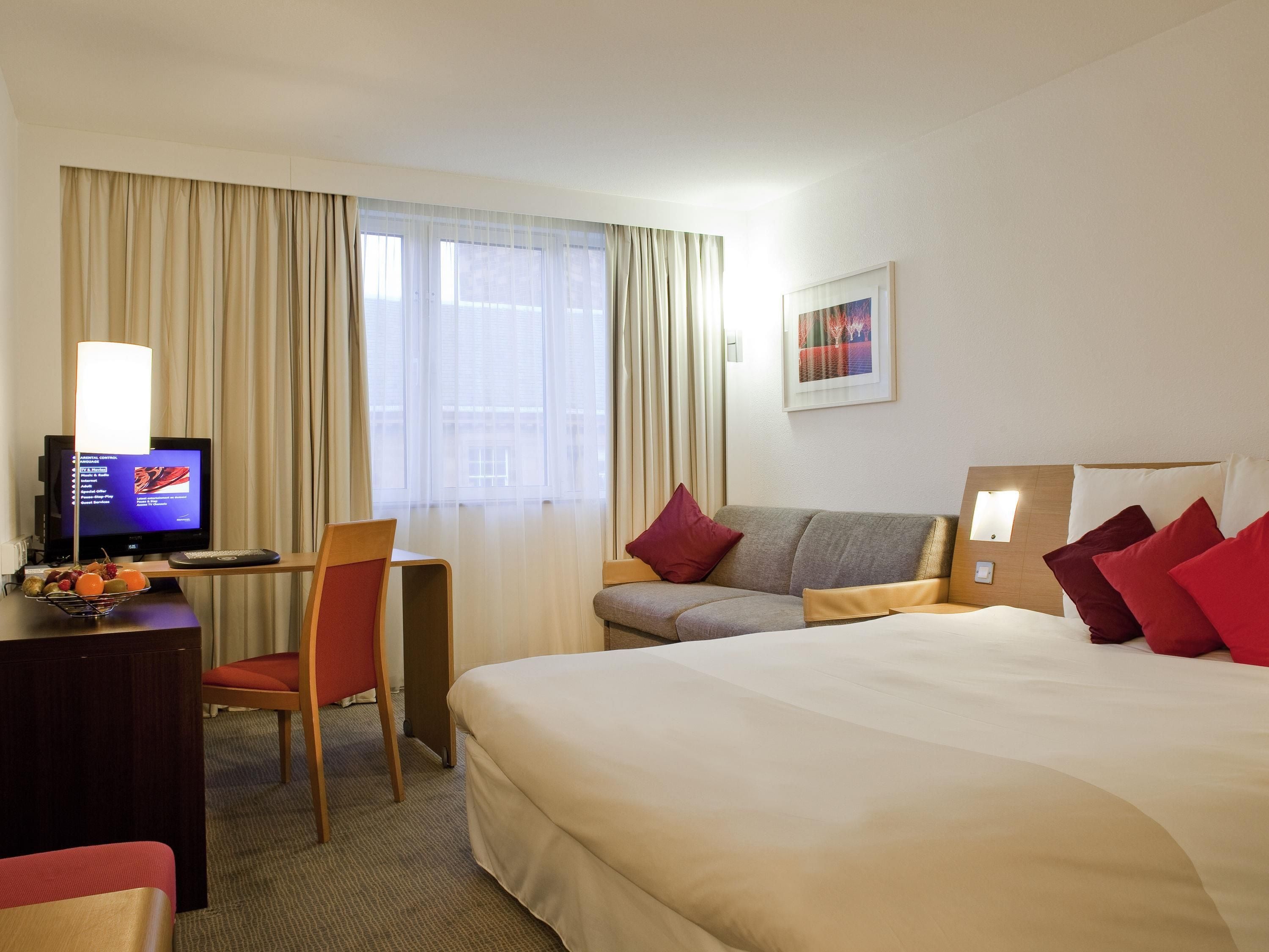 A hotel room with a bed, a desk with a computer, a chair, a television, and a window with curtains.