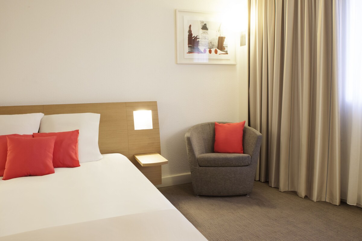 A hotel room with a bed, a nightstand, a chair, and a window with curtains.