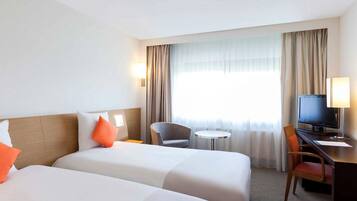Habitación de hotel con dos camas, un escritorio con televisor, una silla, una mesita y una ventana con cortinas.