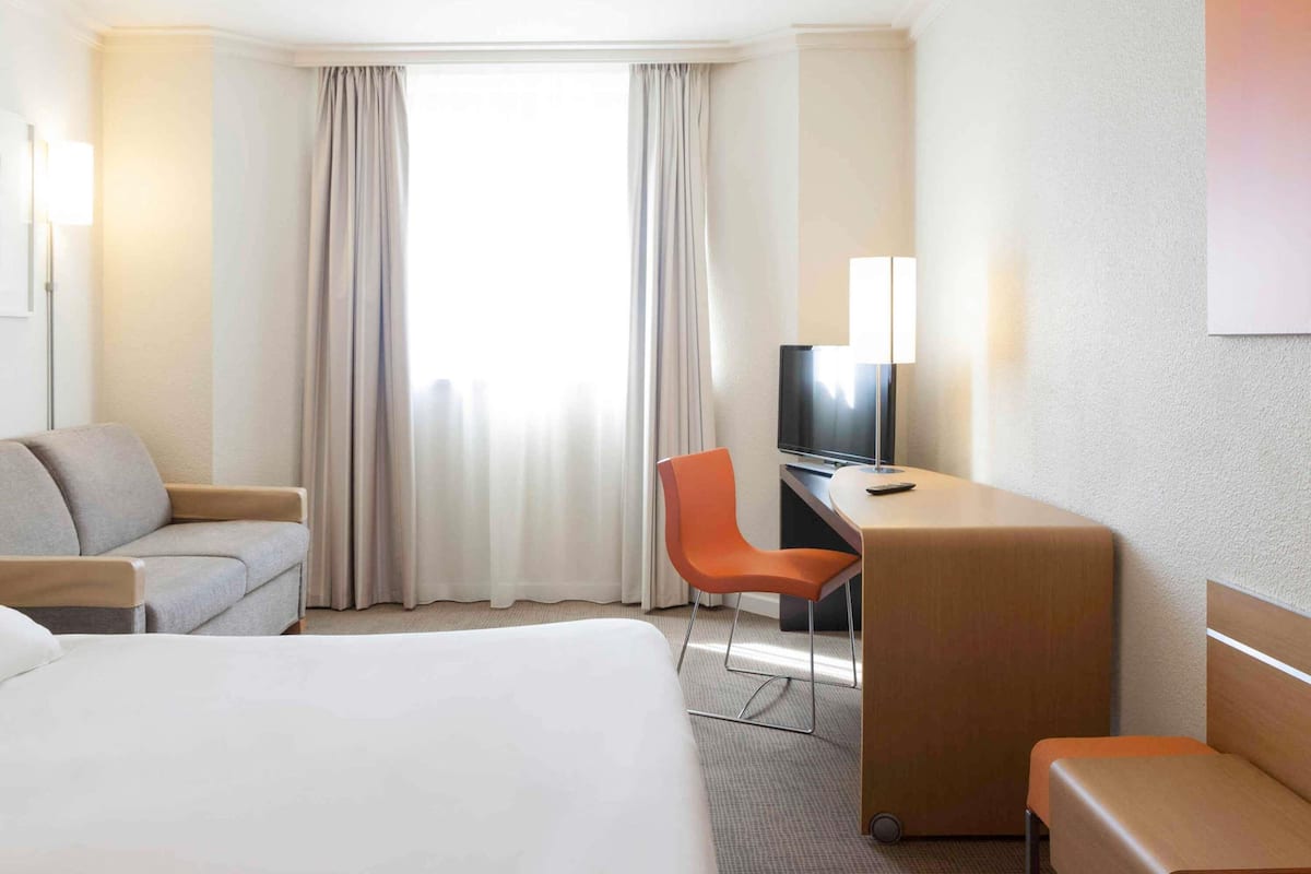 A hotel room with a bed, a desk with a TV, an orange chair, and a large window with curtains.