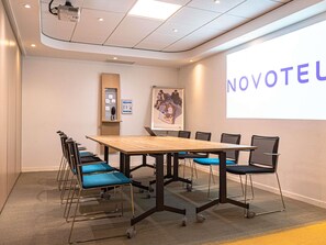 A conference room with a long table, chairs, a projector, and a screen displaying 'NOVOTEL'.