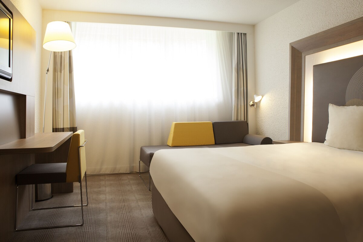 A hotel room with a bed, a desk, a chair, a lamp, and a window with curtains.