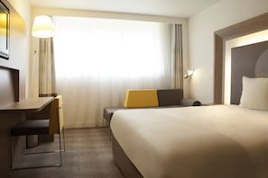 A hotel room with a bed, a desk, a chair, a lamp, and a window with curtains. - Novotel Paris Pont De Sevres (Sevres)