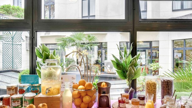 A breakfast buffet with various food items and beverages, including pastries, fruits, and tea, displayed on a pink table in a modern setting with large windows. - Novotel Paris Pont De Sevres (Sevres)