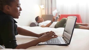 A person using a laptop on a bed in a hotel room with another person reading a book in the background. - Novotel Paris Pont De Sevres (Sevres)