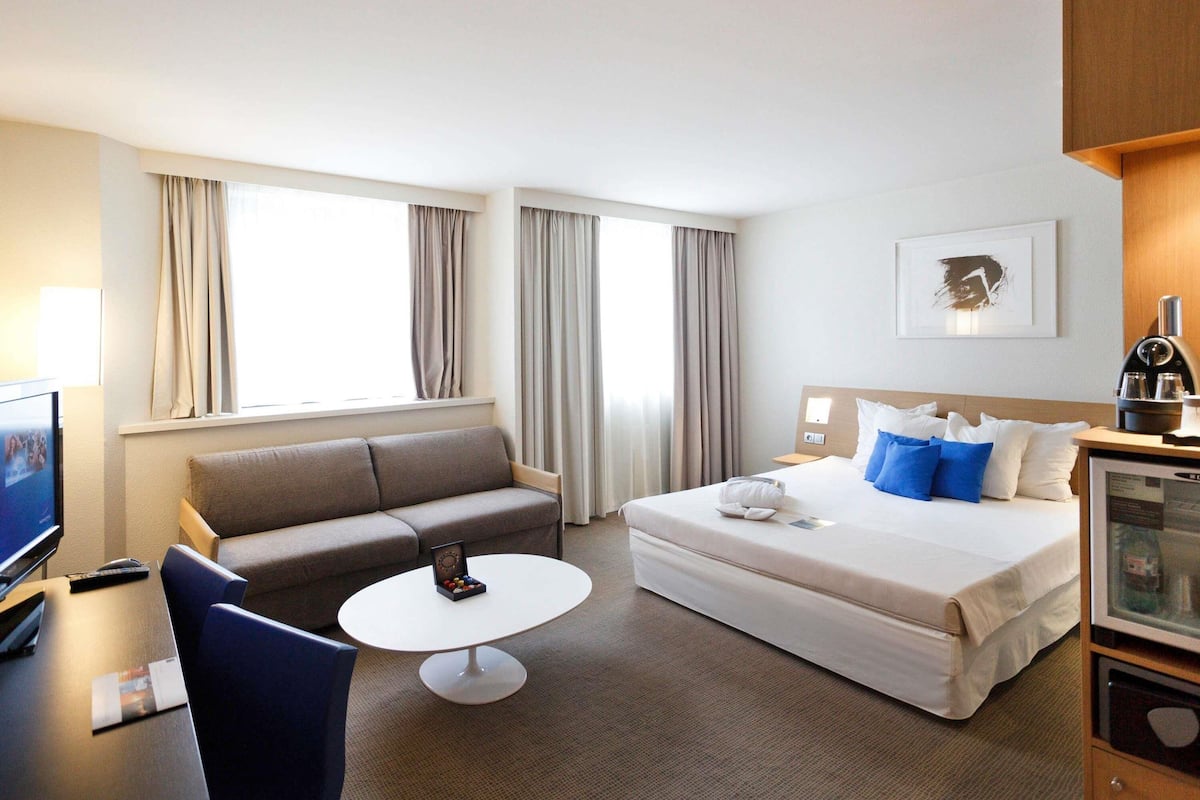 A hotel room with a bed, a sofa, a small round table, a TV, and a window with curtains.