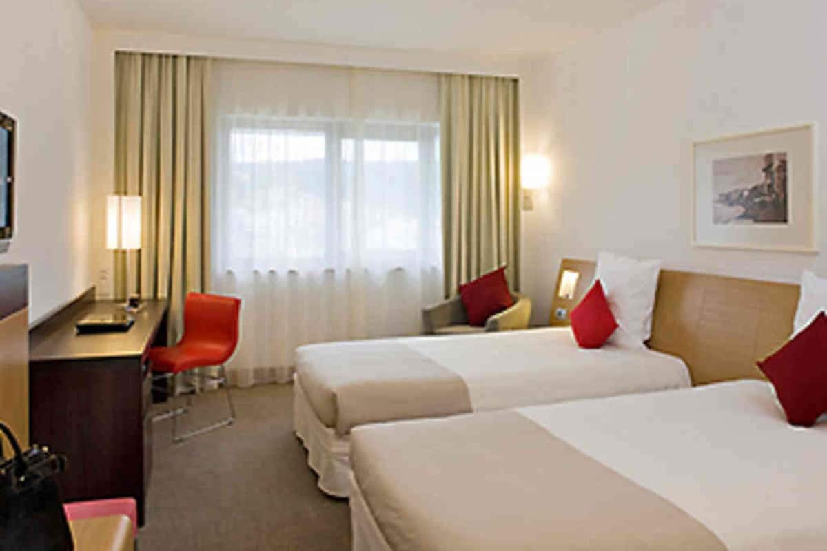 A hotel room with two beds, a desk, a chair, a lamp, and a window with curtains.
