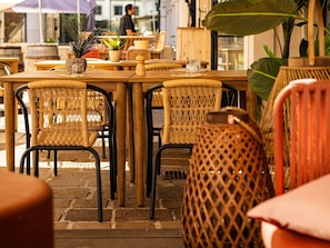 Un restaurant avec des chaises et des tables en osier, des plantes en pot, et une personne qui marche au loin.