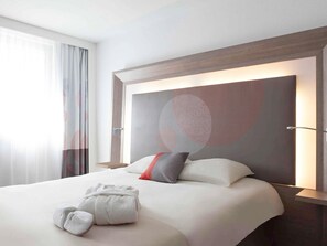 A hotel room with a large bed, a headboard with a geometric design, a bedside table, and a window with sheer curtains.
