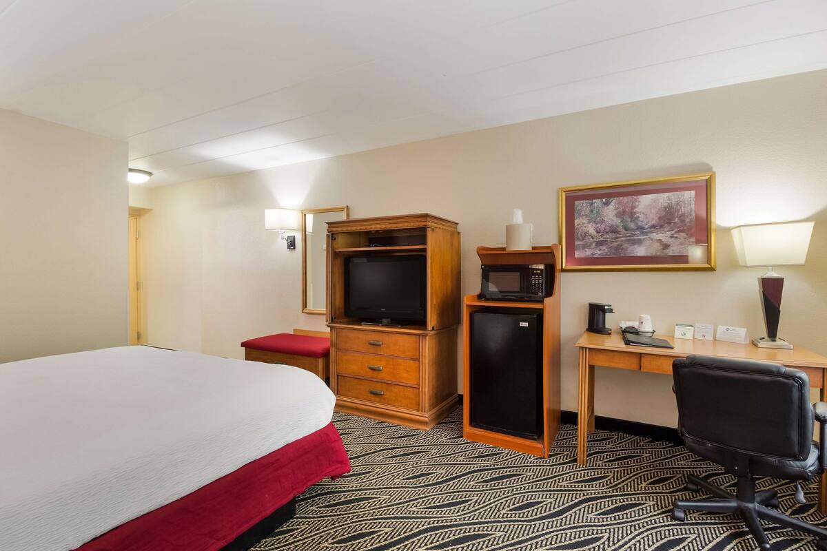 Quarto de hotel com cama, cómoda de madeira, secretária com computador, micro-ondas e pequeno frigorífico.
