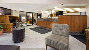 A reception area with a wooden desk, a patterned chair, a black metal trash bin, and a reception counter. - Best Western Braddock Inn (Cumberland)