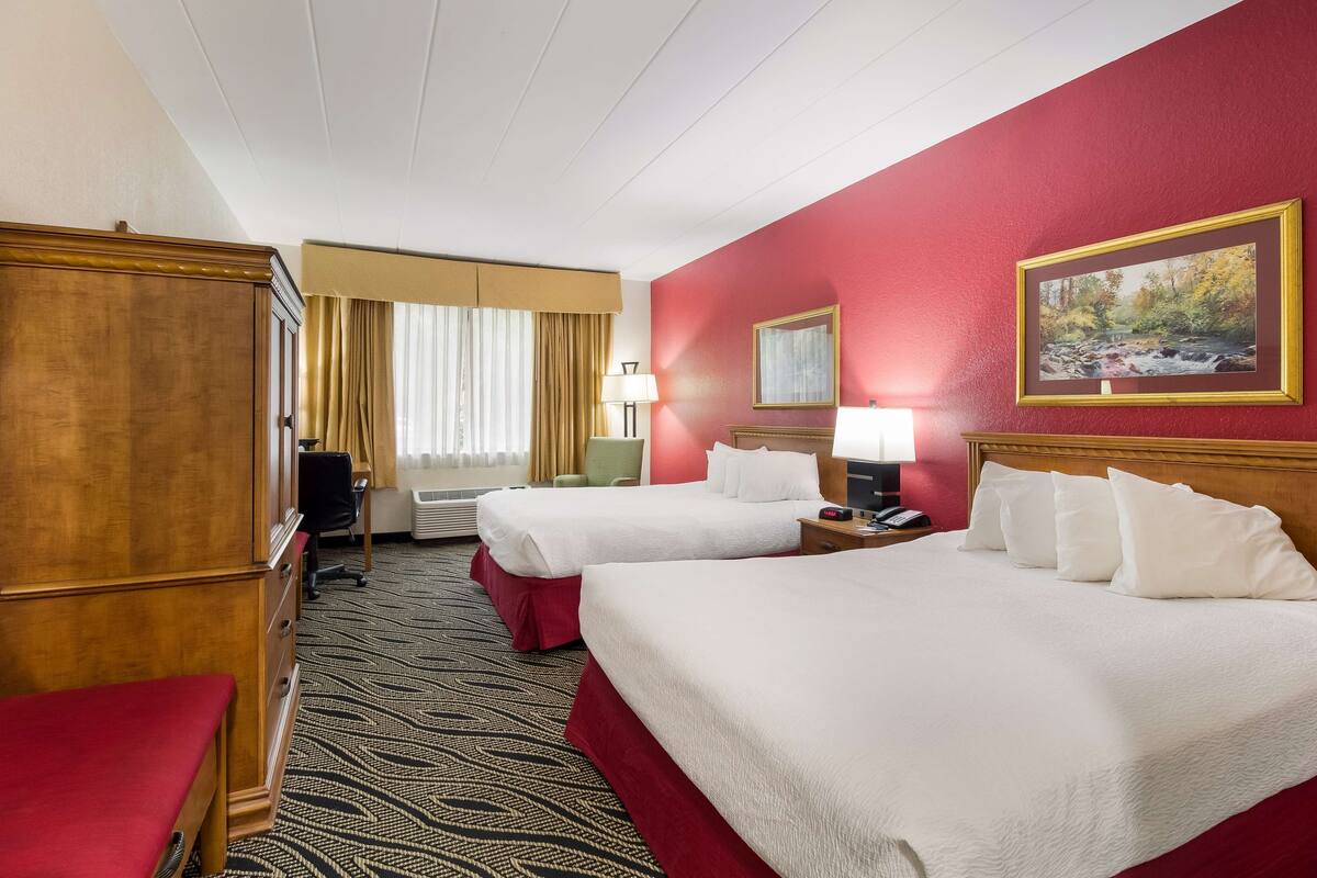 Quarto de hotel com duas camas, um guarda-roupa de madeira, uma escrivaninha, uma cadeira e uma janela com cortinas.