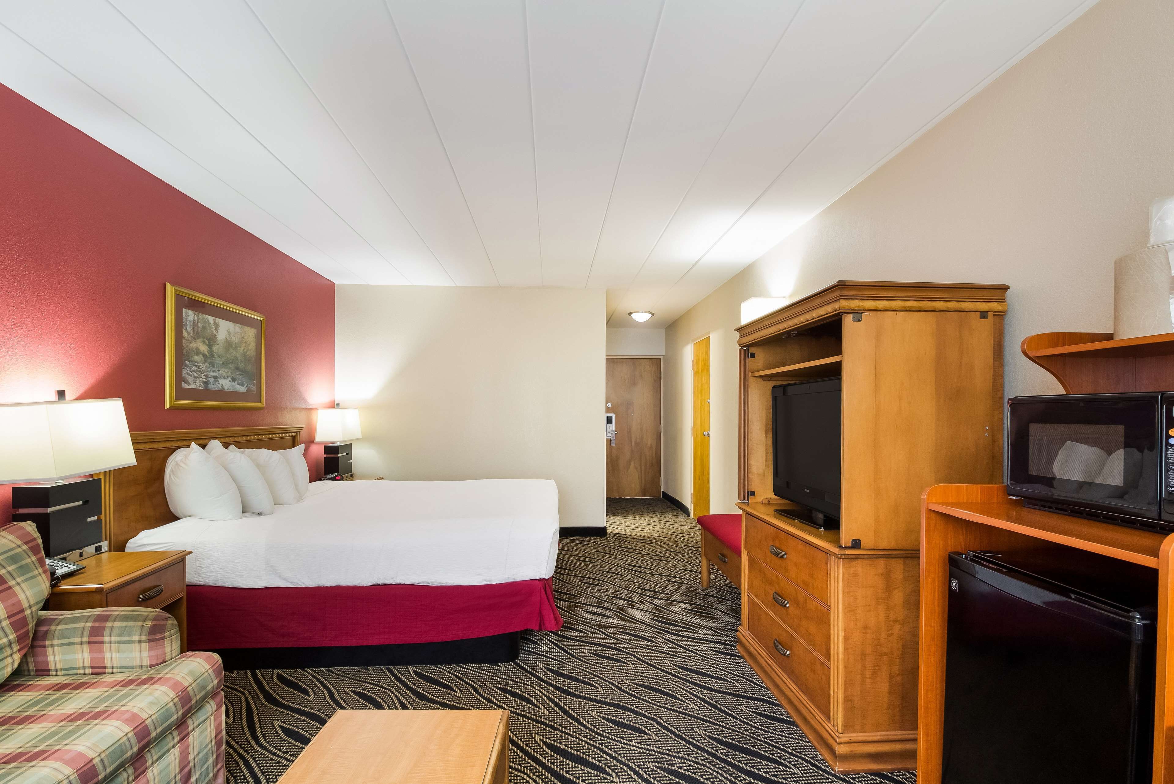 Een hotelkamer met een bed, een televisie, een koelkast, een houten kast, een bureau, een stoel en een schilderij aan de muur.