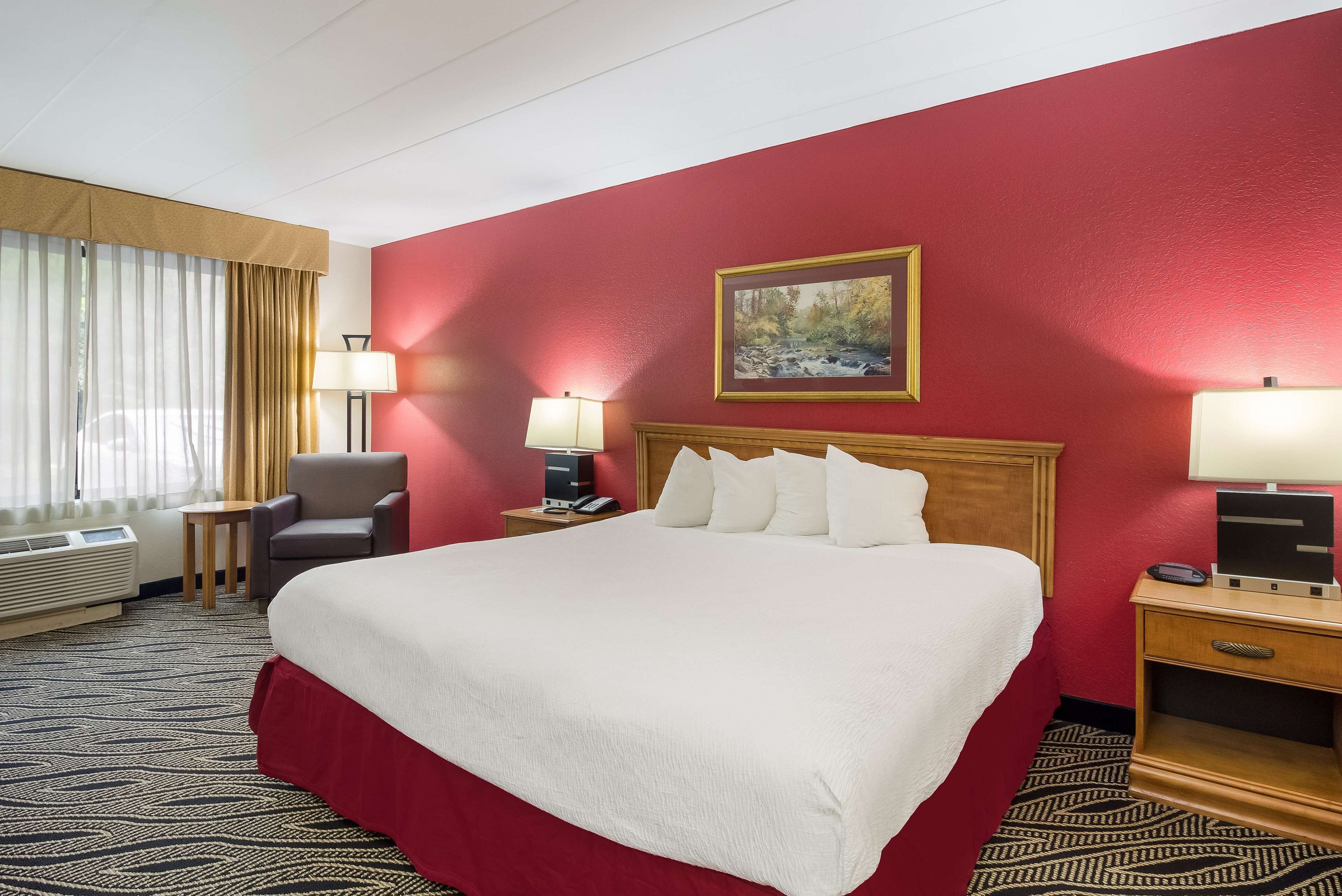 Quarto de hotel com uma cama grande, uma escrivaninha com luminária, uma cadeira, um quadro na parede e uma janela com cortinas.