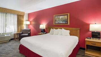 Quarto de hotel com uma cama grande, uma escrivaninha com luminária, uma cadeira, um quadro na parede e uma janela com cortinas.