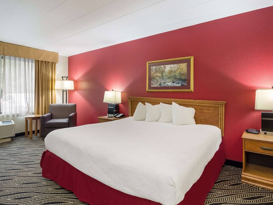 Quarto de hotel com uma cama grande, uma escrivaninha com luminária, uma cadeira, um quadro na parede e uma janela com cortinas.
