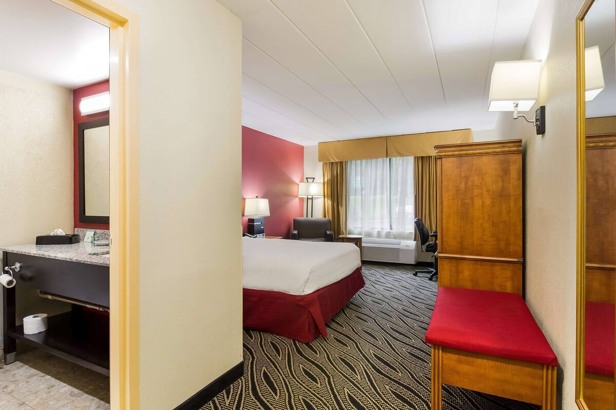 Quarto de hotel com cama, escrivaninha, cadeira, espelho, guarda-roupa e banheiro com vaso sanitário e pia.