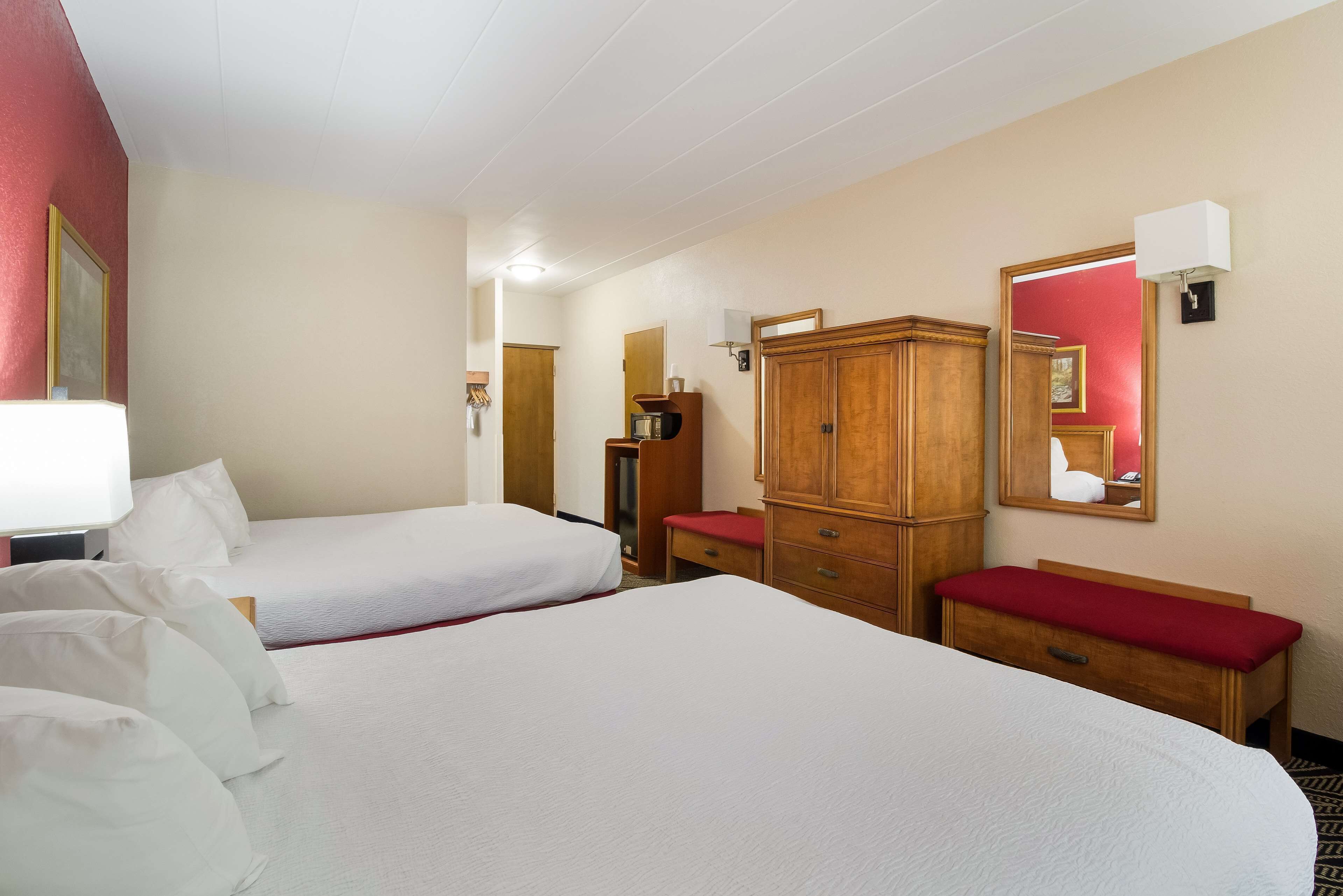 A hotel room with two beds, a wooden wardrobe, a mirror, and a red bench.