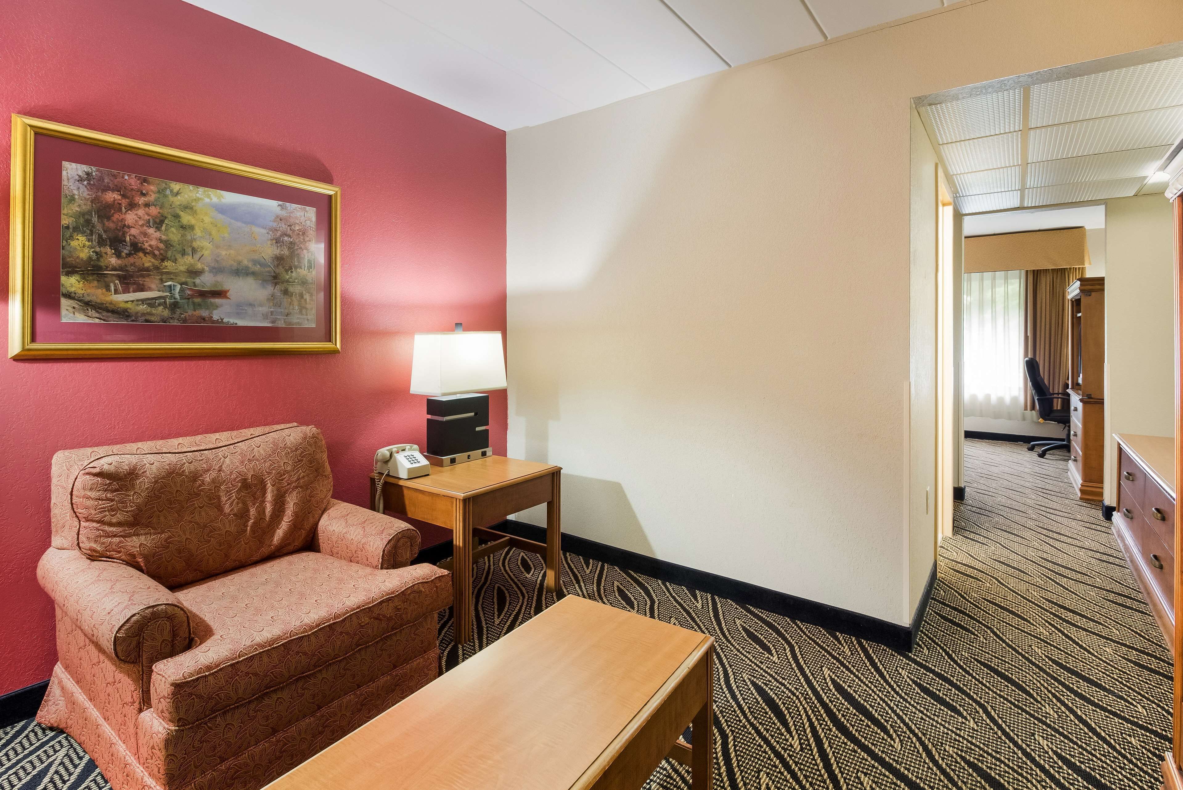 A hotel room with a red wall, a painting, a wooden armchair, a small table, a lamp, and a carpeted floor.