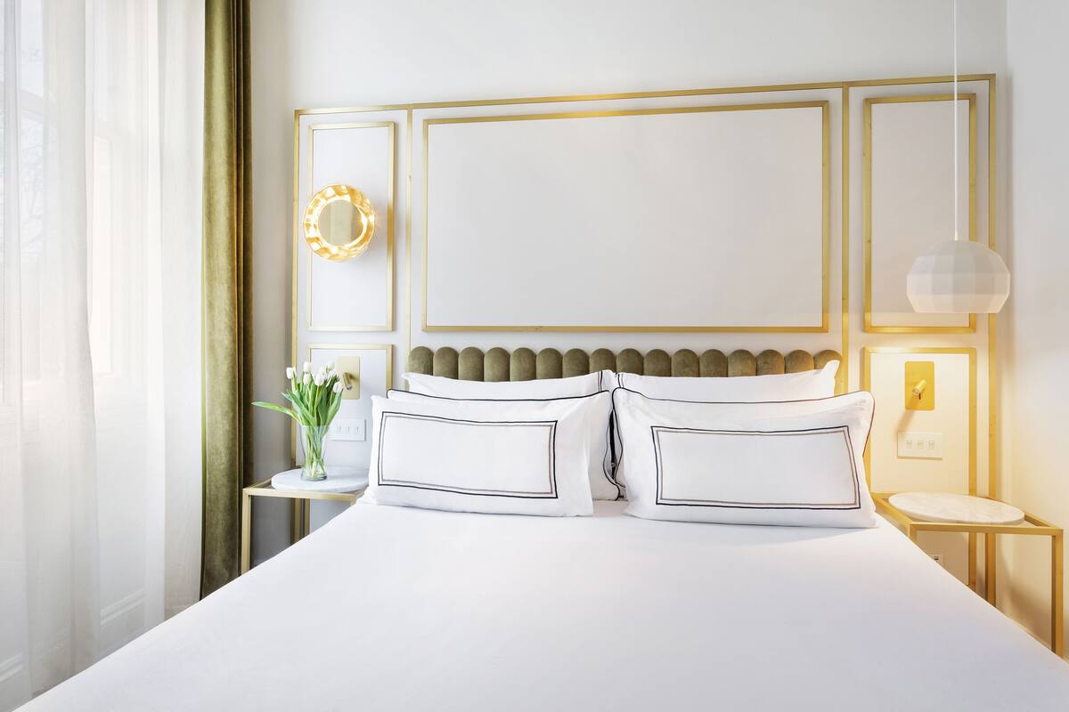 Ein ordentlich eingerichtetes Hotelzimmer mit einem großen Bett, einem Nachttisch mit Blumenvase, einem Wandspiegel und einer Pendelleuchte.