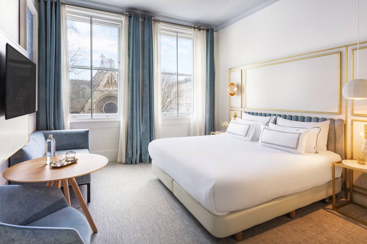 Ein Hotelzimmer mit einem großen Bett, einem runden Holztisch mit zwei Stühlen, einem an der Wand befestigten Fernseher und einem Fenster mit Vorhängen.