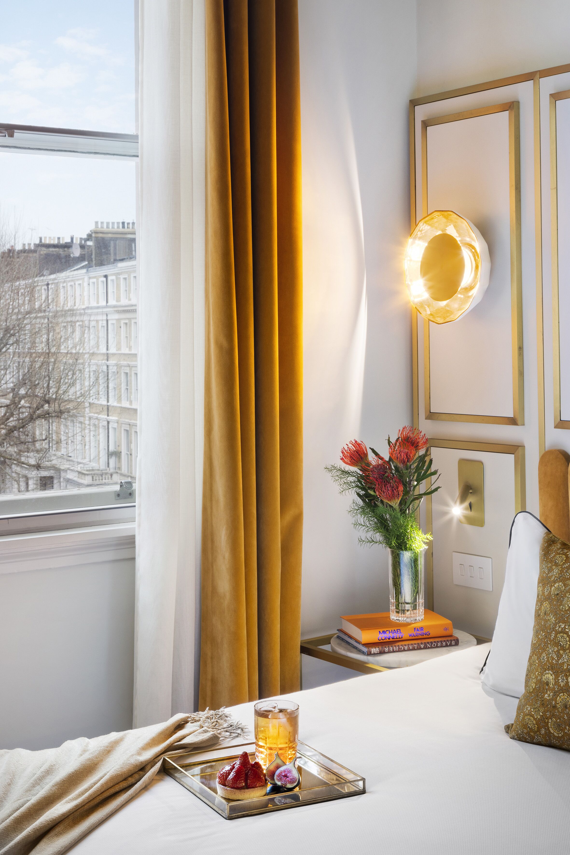 Une chambre d’hôtel avec un lit, une table de chevet ornée d’un vase de fleurs, un plateau avec des fruits et une boisson, et une vue sur les bâtiments à travers la fenêtre.